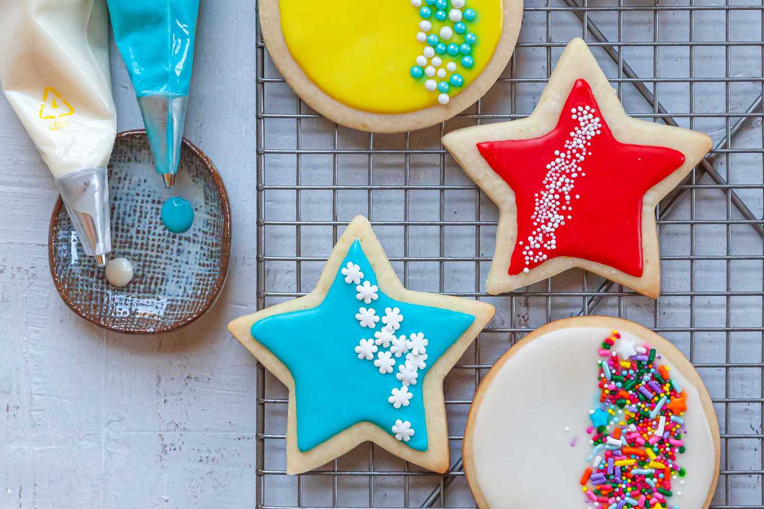 Two piping bags filled with Royal icing are set next to a cooling rack with decorated sugar cookies on it.