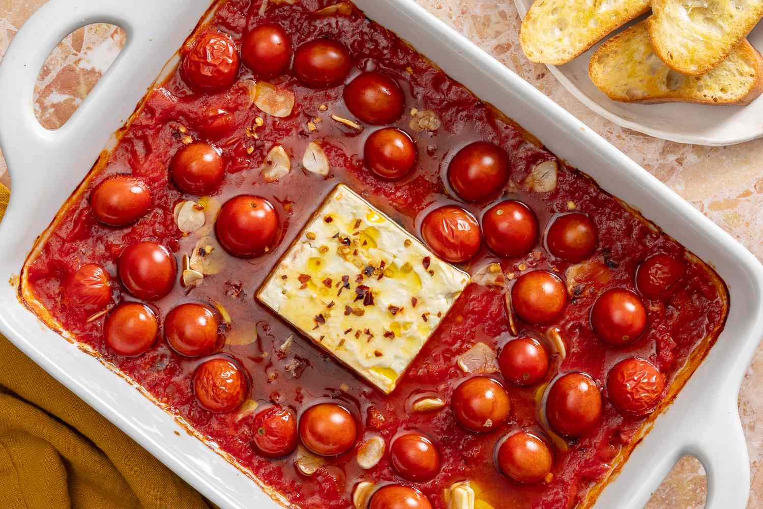 Overhead view of a white baking dish with garlic, tomatoes, cannellini beans and a block of feta covered in olive oil and red pepper flakes next to a plate of buttered french bread slices and a napkin