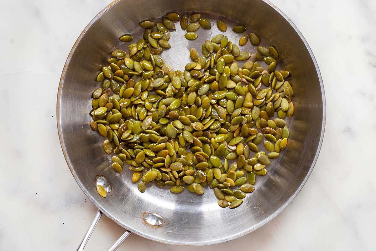 Silver saute pan on a marble background with raw pumpkin seeds inside.
