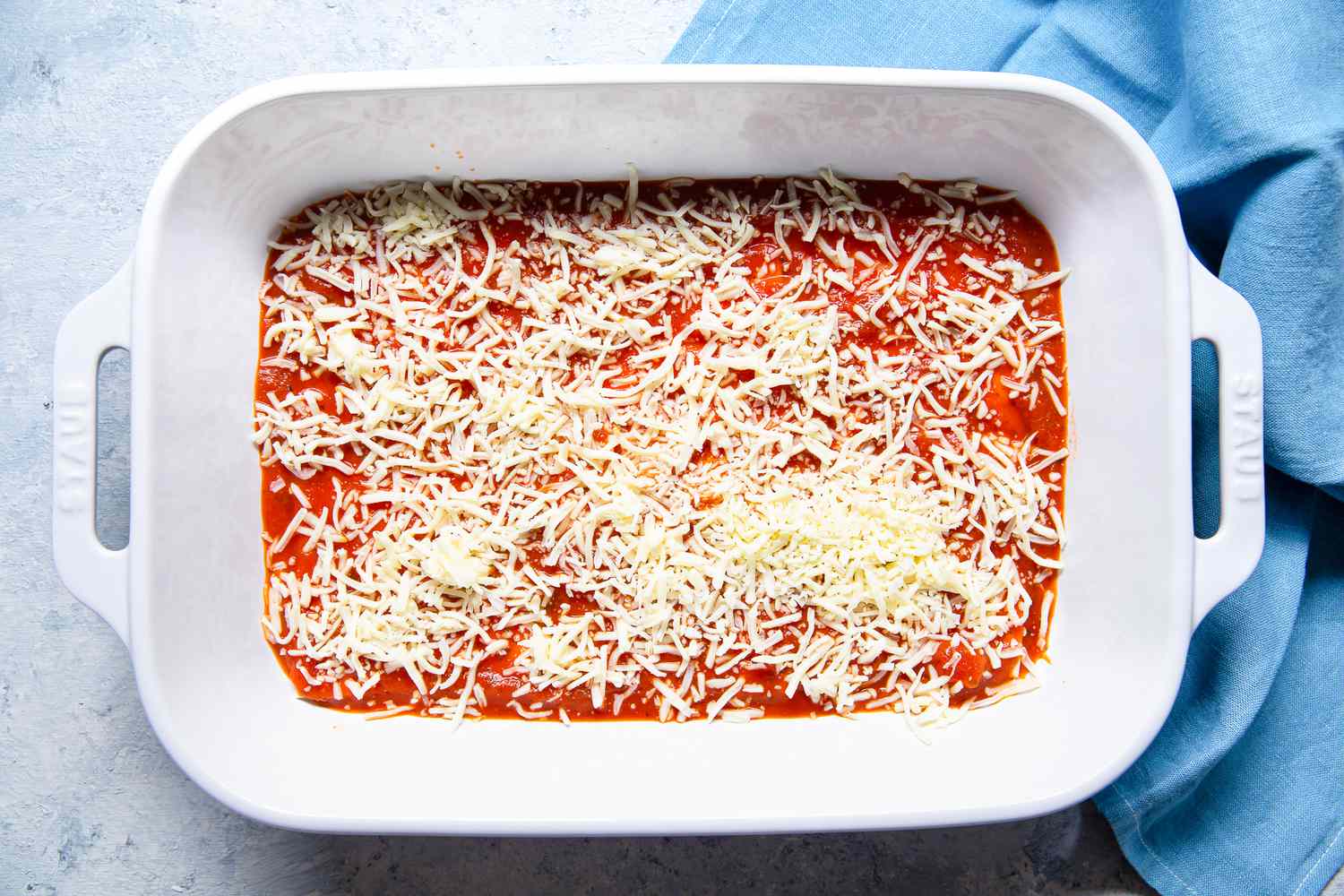 Shredded Cheese Added in the Casserole Dish Next to the Sky Blue Kitchen Towel for Pepperoni Pizza Lasagna