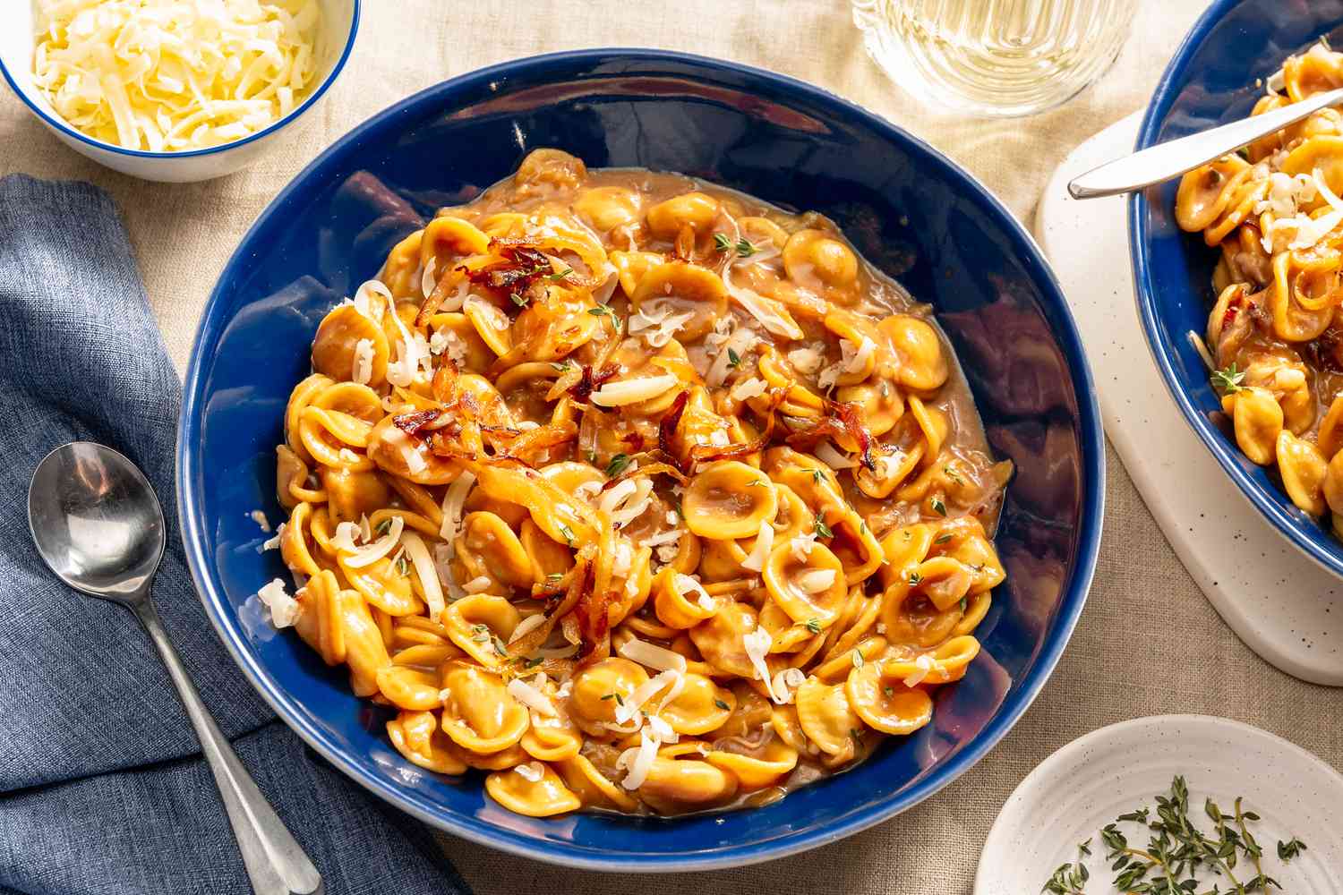 servings of French onion pasta in two bowls at a table setting with a small bowl of grated cheese, another bowl with fresh thyme, and a glass of water 