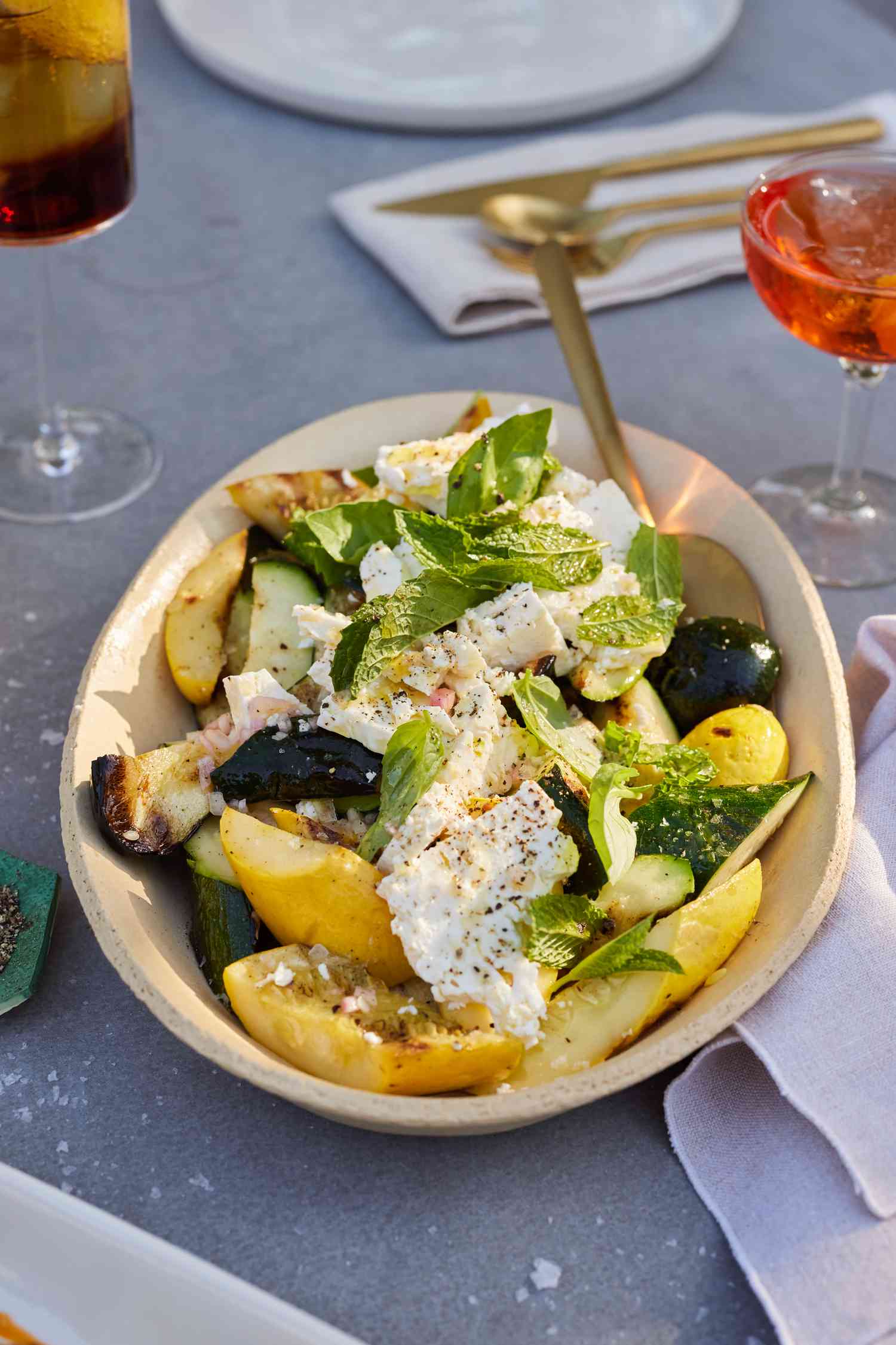 Bowl of Grilled Zucchini and Yellow Squash with Champagne Vinaigrette with a Spoon Surrounded by Drinks and Other Utensils on a Table
