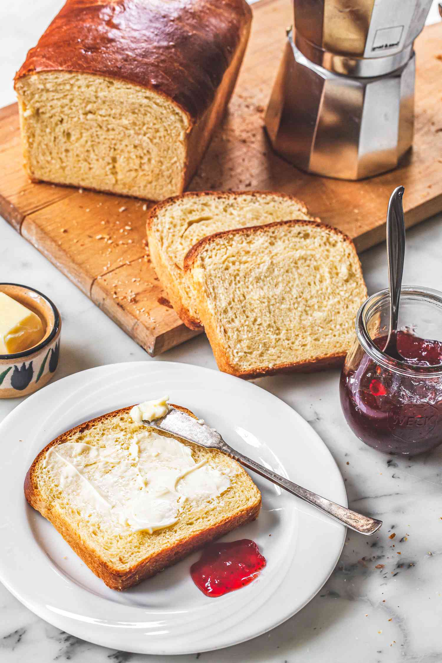 Brioche Sandwich Loaf on a Wooden Cutting Board with Slices Cascading Off onto the Counter Next to a Plate with a Dollop of Jam and a Slice Smeared in Butter. In the Surroundings, a Moka Pot on the Cutting Board, a Jar of Jam on the Counter, and a Dish of Butter