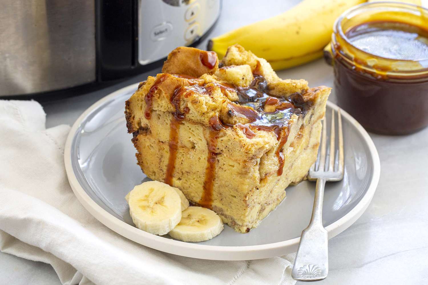 Slice of banan bread pudding drizzeld with caramel saucemade in the slow cooker on a gray plate with a fork and sliced bananas. The slow cooker is in the background.