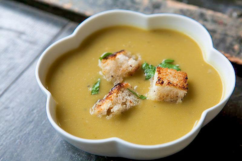 A close-up of a bowl of split pea soup with croutons and chopped herbs on top