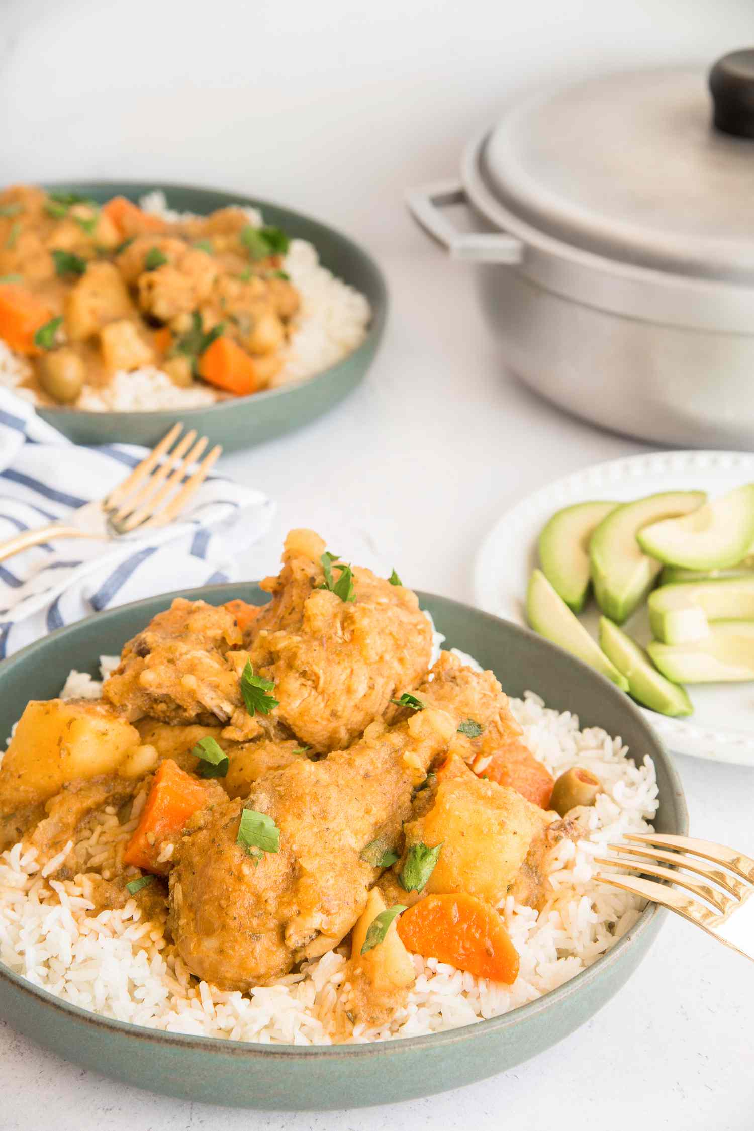Bowl of Pollo Guisado Served Over Rice, and in the Surroundings, a Plate with Avocado, Another Bowl of Pollo Guisado, and a Caldero 