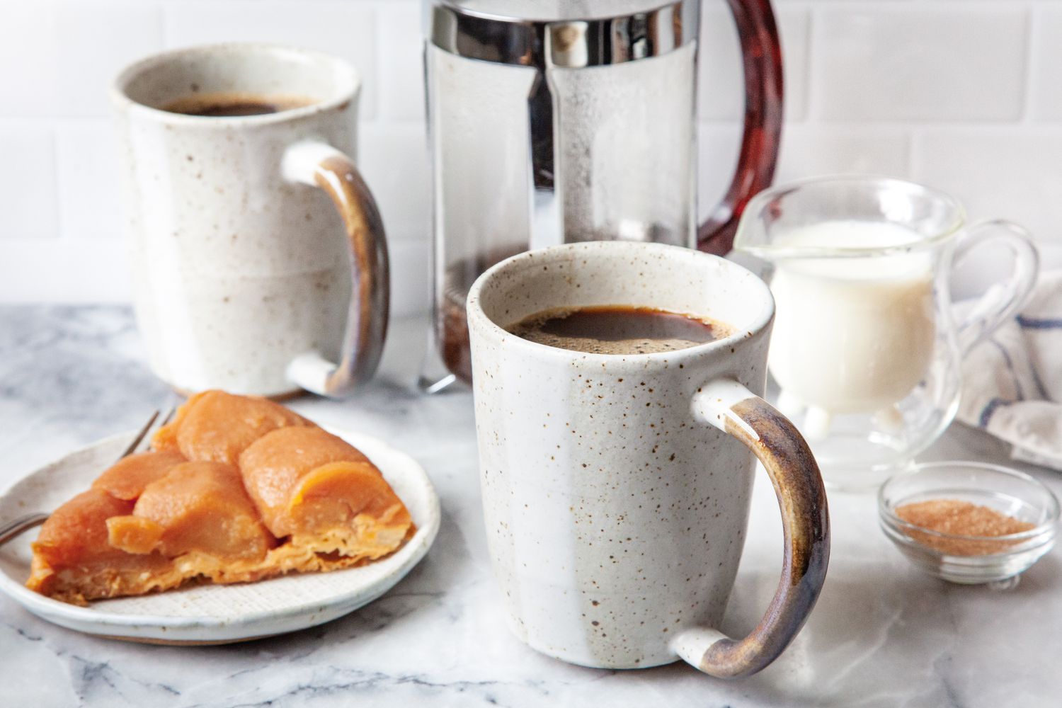 Cup of French Press Coffee Surrounded by a Plate with a Breakfast Pastry, a Bowl of Sugar, a Small Pitcher of Milk, Another Cup of Coffee, and a French Press