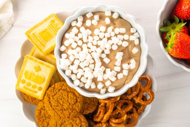 A bowl of hot cocoa dip topped with marshmallows surrounded by cookies pretzels and crackers on a plate