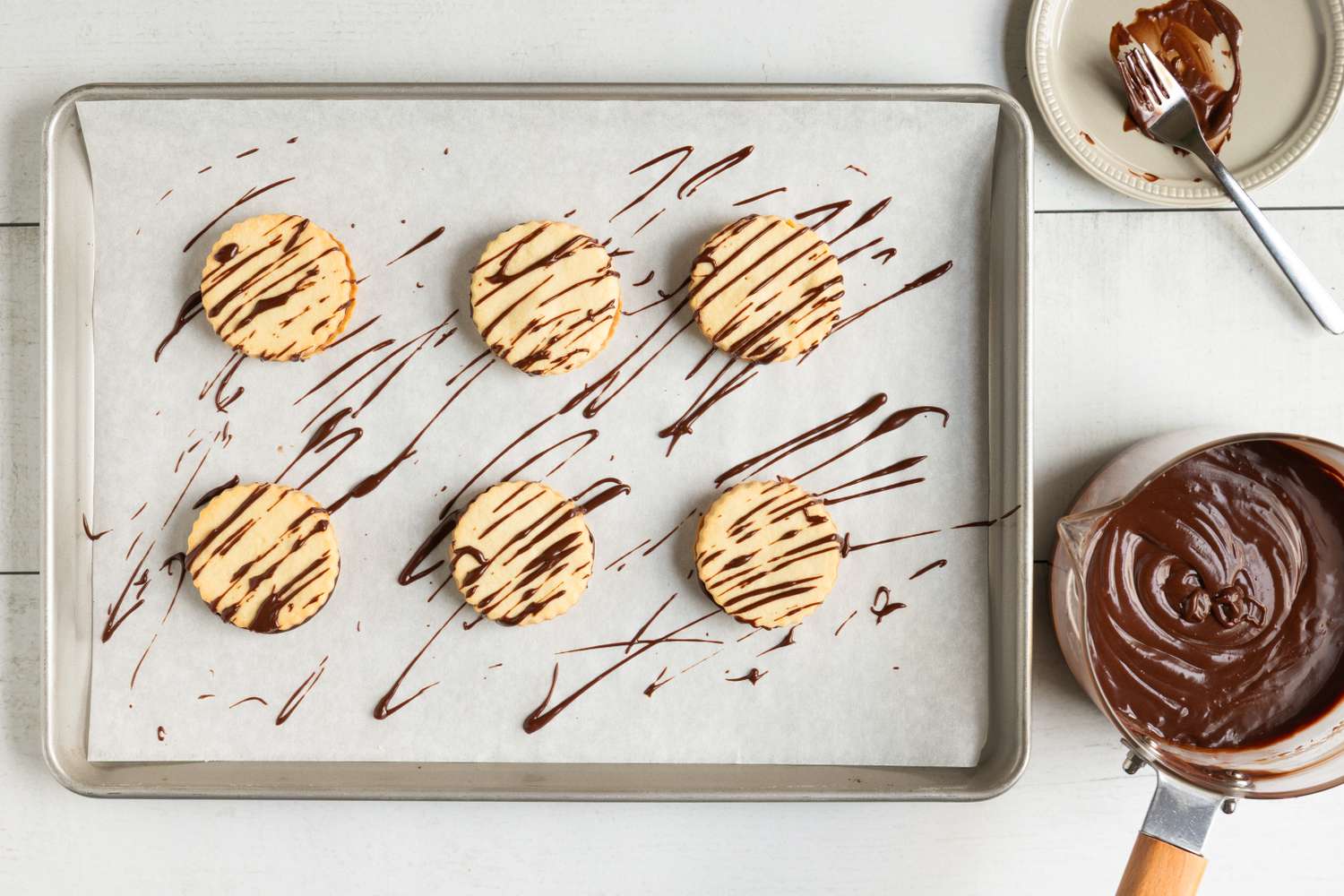 Cookies on a baking sheet with chocolate drizzle a bowl of melted chocolate and a small plate with a fork