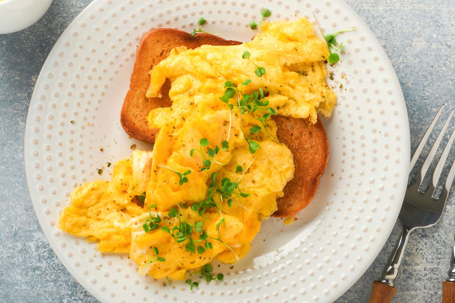 Overhead view of a white plate of a pieced of toast with scrambled eggs on top next to a fork