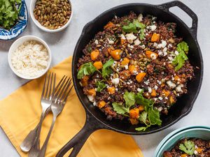 Horizontal view of a large cast iron skillet filled with a butternut squash skillet dinner. Cilantro, quinoa, butternut squash and cotija cheese are visible in the pan. To the left is a yellow napkin with three forks on it. Above the forks are three small bowls. One small bowl of rice, a ramiken of roasted pumpkin seeds and a small platter of cilantro.
