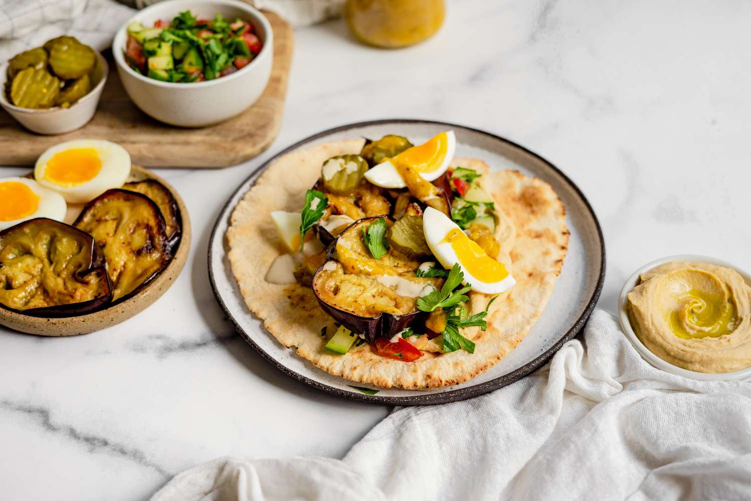 Sabich on a Plate Surrounded by Bowl of Hummus, Bowl of Israeli Salad, and a Bowl of Eggplant and Boiled Eggs for Sabich