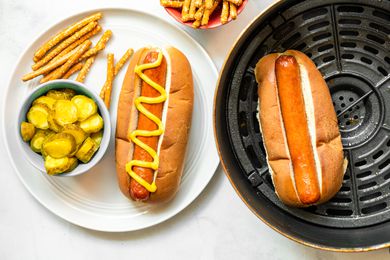 A Plate of Air Fryer Hot Dog in a Bun Topped with Mustard and a Side of Pickles and Pretzels. Next to It Is the Air Fryer Basin with Another Hot Dog in a Bun and a Bowl of Pretzels.