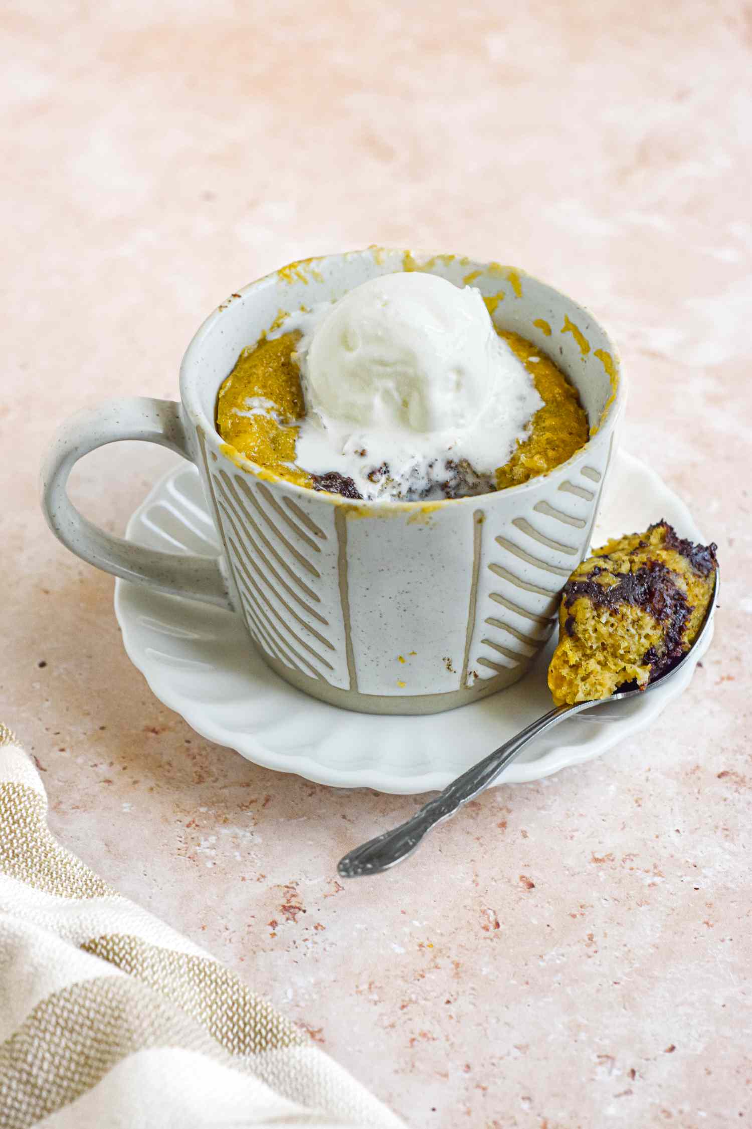 Mug of Banana Bread Chocolate Chunk Mug Cake Topped with a Dollop of Ice Cream and Spoonful of Cake Resting on a Saucer (Mug on the Saucer Next to the Spoon)