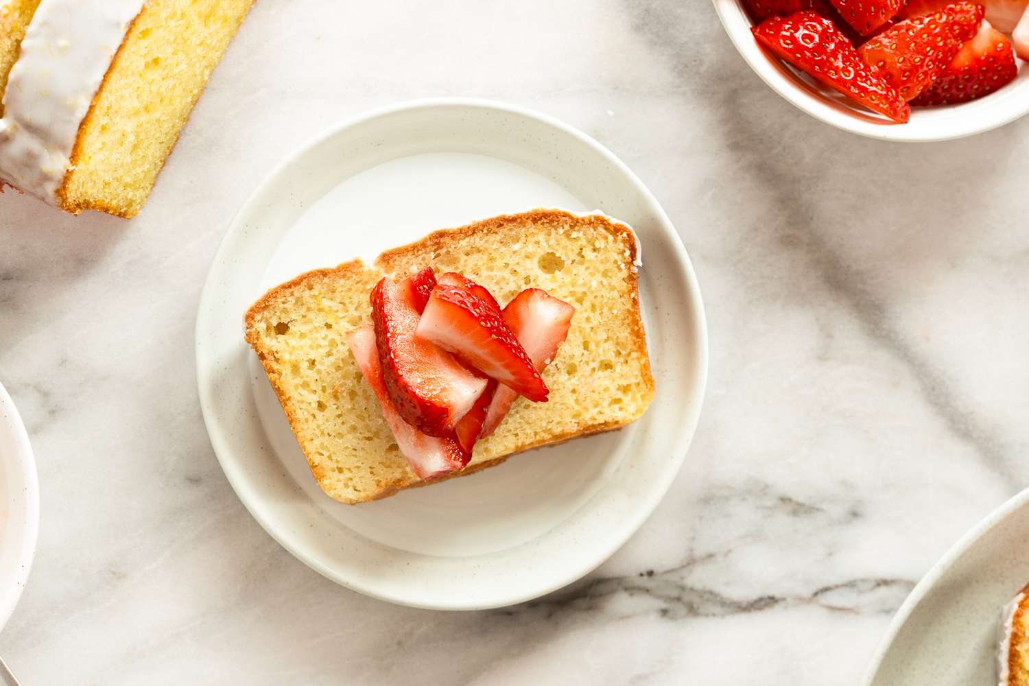Slice of pound cake topped with sliced strawberries on a small white plate, surrounded by other dessert components.