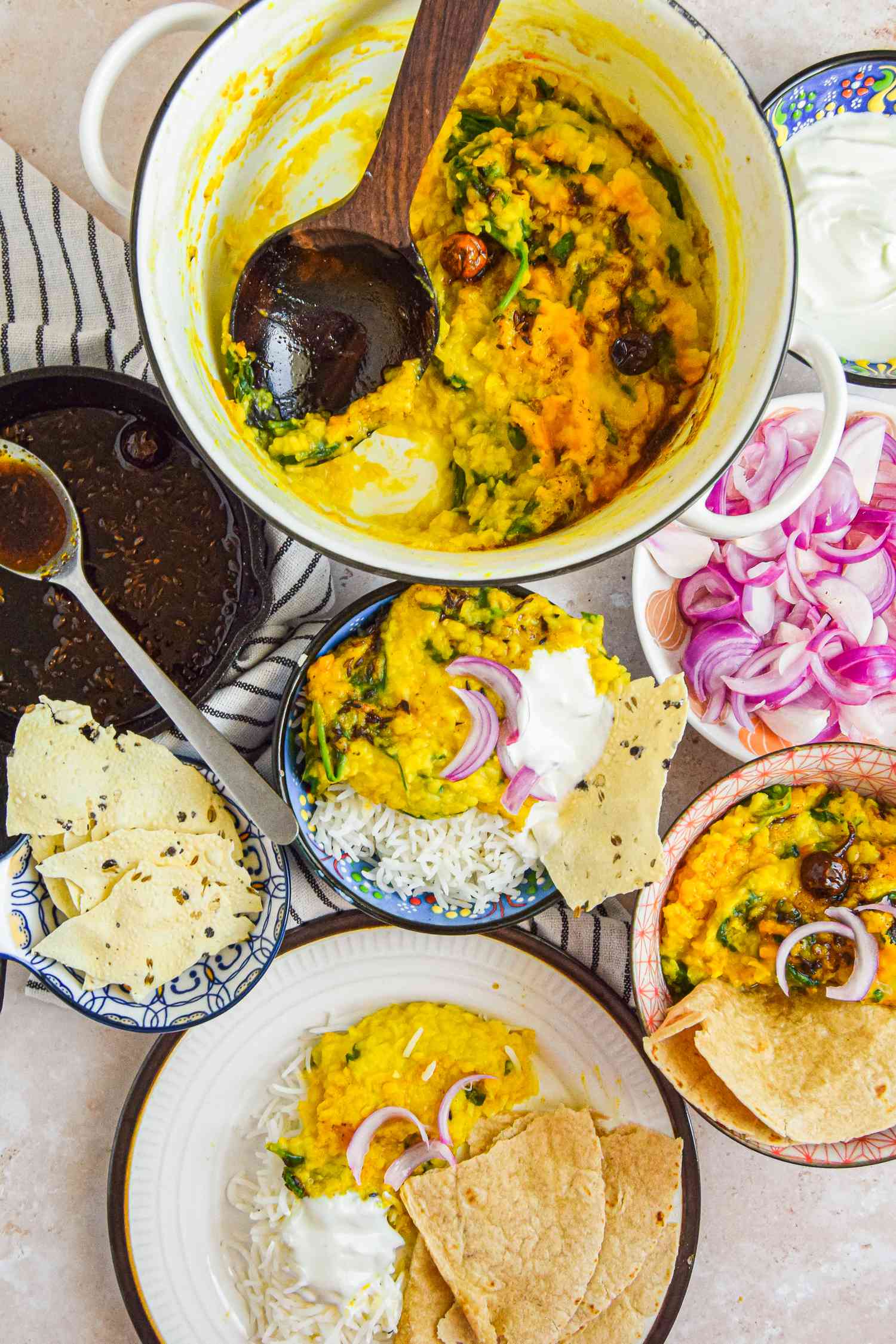 Table Setting with Daal Palak in a Dutch Oven, Two Servings of Daal Palak in Two Bowl with Rice and Papad, a Plate with Another Serving on Rice with a Piece of Roti, a Small Cast Iron Skillet with Leftover Tadka, and a Bowl of Papad