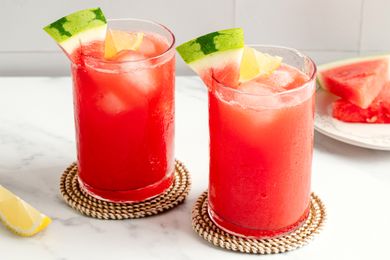Two glasses of watermelemonade with ice and fruit garnishes placed on coasters on a surface