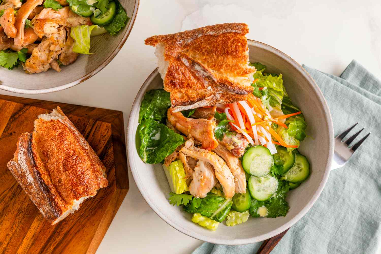 Overhead shot: banh mi salad bowl with a baguette piece next to a wooden board with another baguette piece, a table napkin with a fork, and another bowl of salad