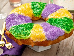 King cake (all decorated) on a cake stand at a table setting with utensils, a purple napkin, wine glasses, and a stack of plates