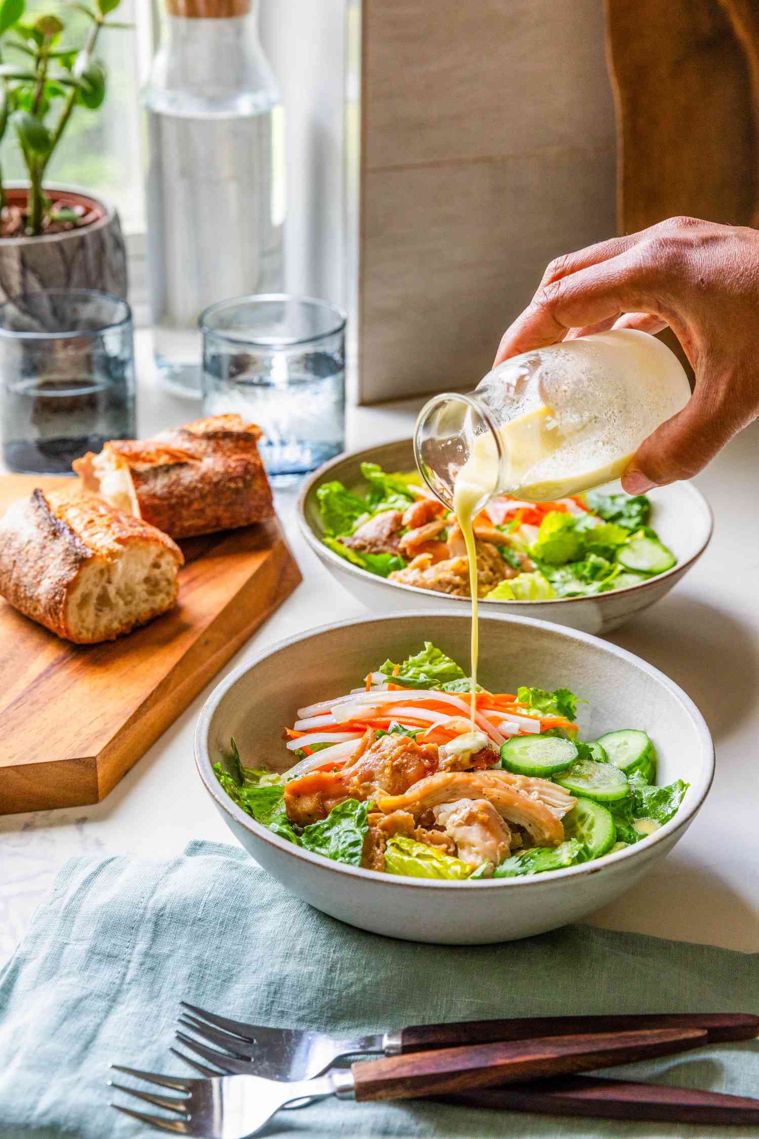 Table setting with bowls of banh mi salad (salad dressing poured onto the most anterior bowl) surrounded by forks on a table napkin, baguette slices on a wooden board, glasses of water, a carafe of water, and a small potted plant