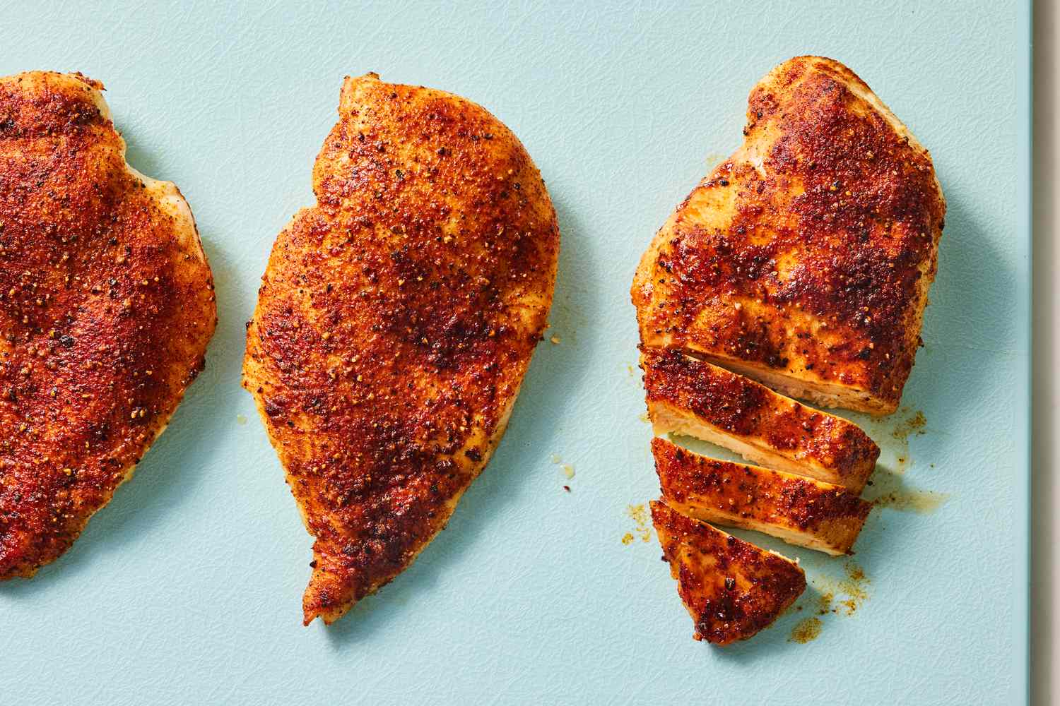 Three seasoned chicken breasts on a surface one sliced into pieces