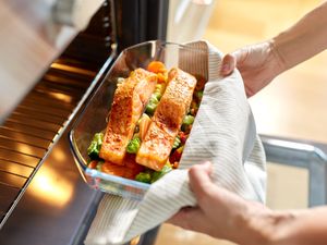 Hands placing a glass dish with roasted salmon and vegetables into an oven
