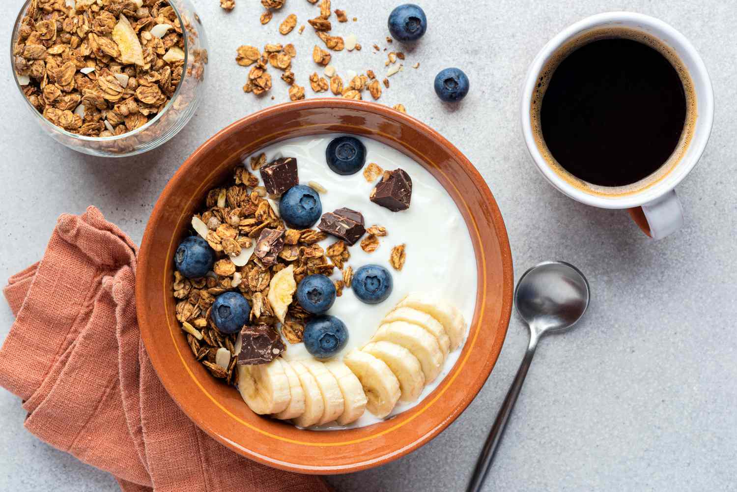 A bowl of yogurt with sliced banana, blueberries, and granola next to a mug of coffee