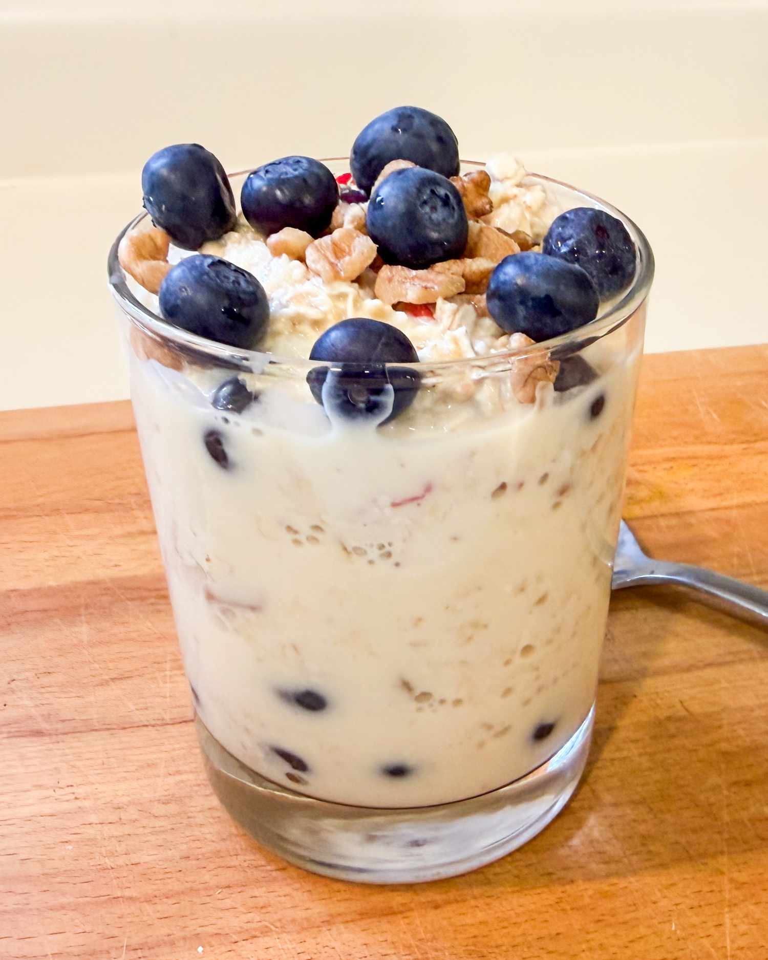 A glass container of oatmeal topped with blueberries and nuts placed on a wooden surface with a spoon nearby