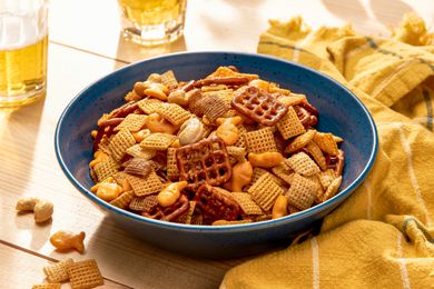 Sweet and spicy chex mix in a bowl at a table setting with two glasses of beer and a yellow kitchen towel