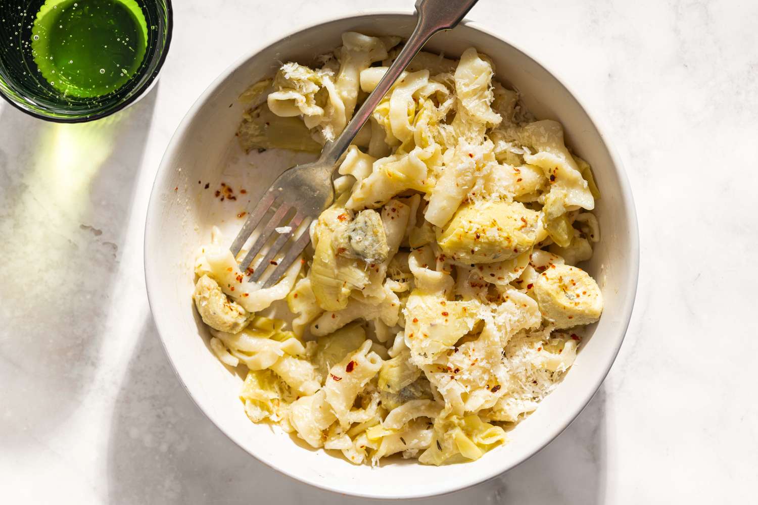 A bowl of pasta with artichokes and lemon garnished with a sprinkle of red pepper flakes and paired with a glass of green liquid