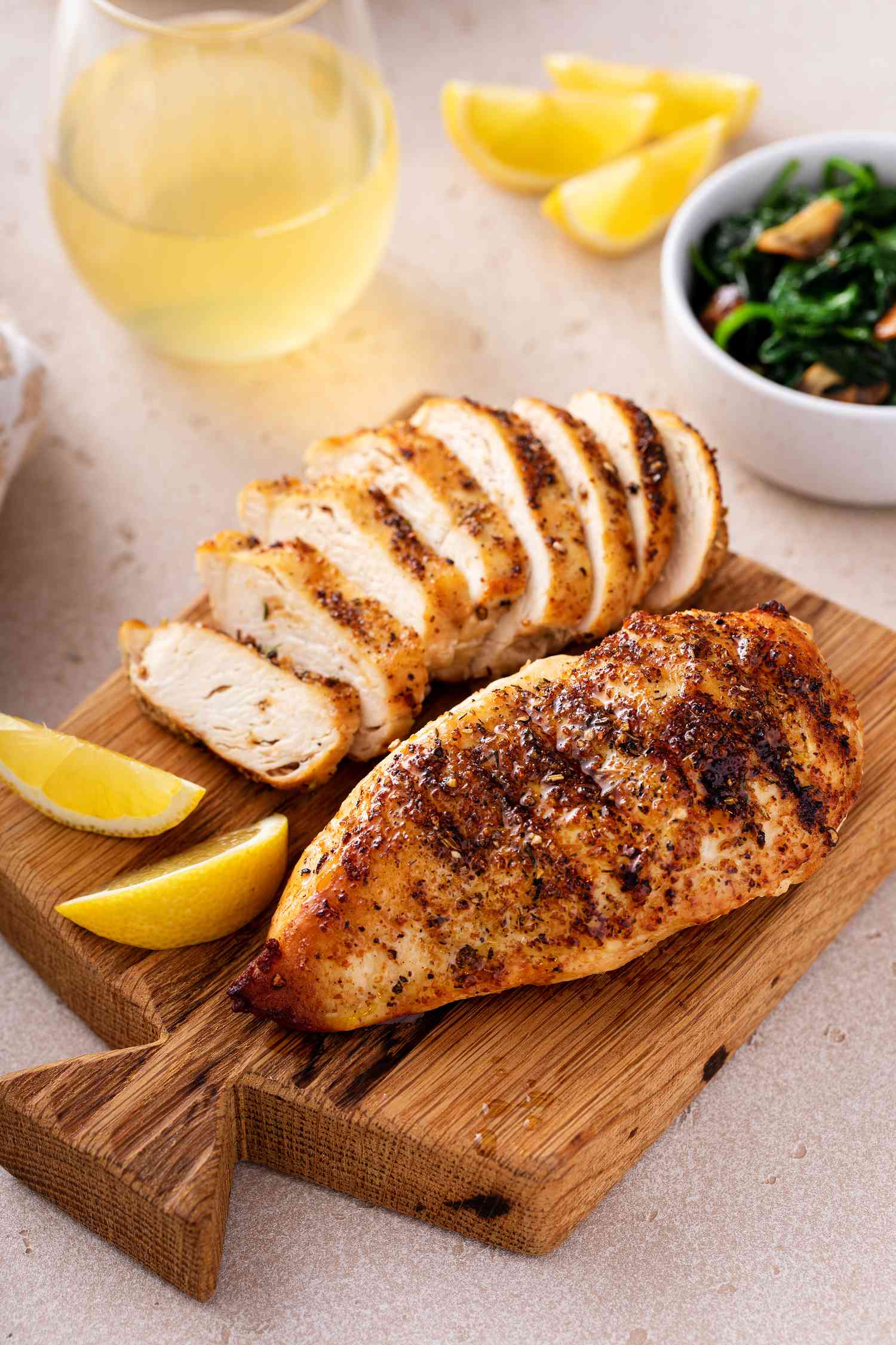 Grilled chicken breasts (one cut into slices, the other whole) on a wooden board with lemon slices, and in the surroundings, a glass of wine, more lemon slices, and a bowl of spinach. 