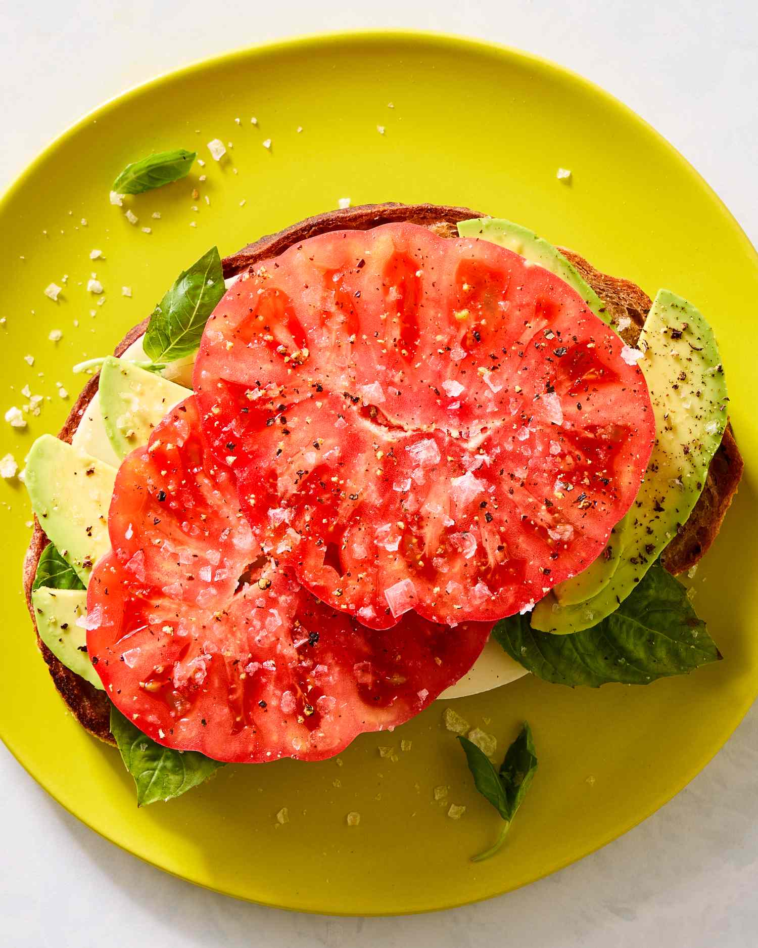 An open sandwich with sliced tomato, avocado, and basil leaves on a yellow plate
