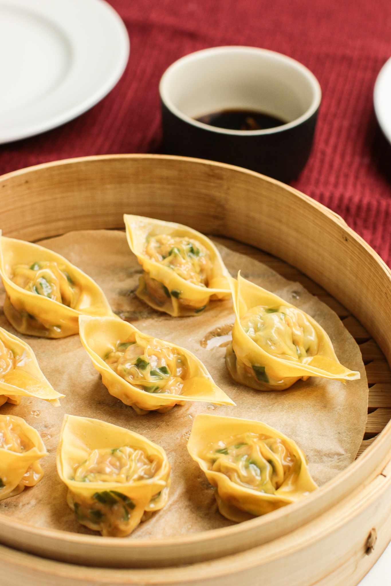 Pork and Chive Dumplings in a Lined Bamboo Steamer Next to a Kitchen Towel with a Bowl of Vinegar Sauce and Two Plates on Top