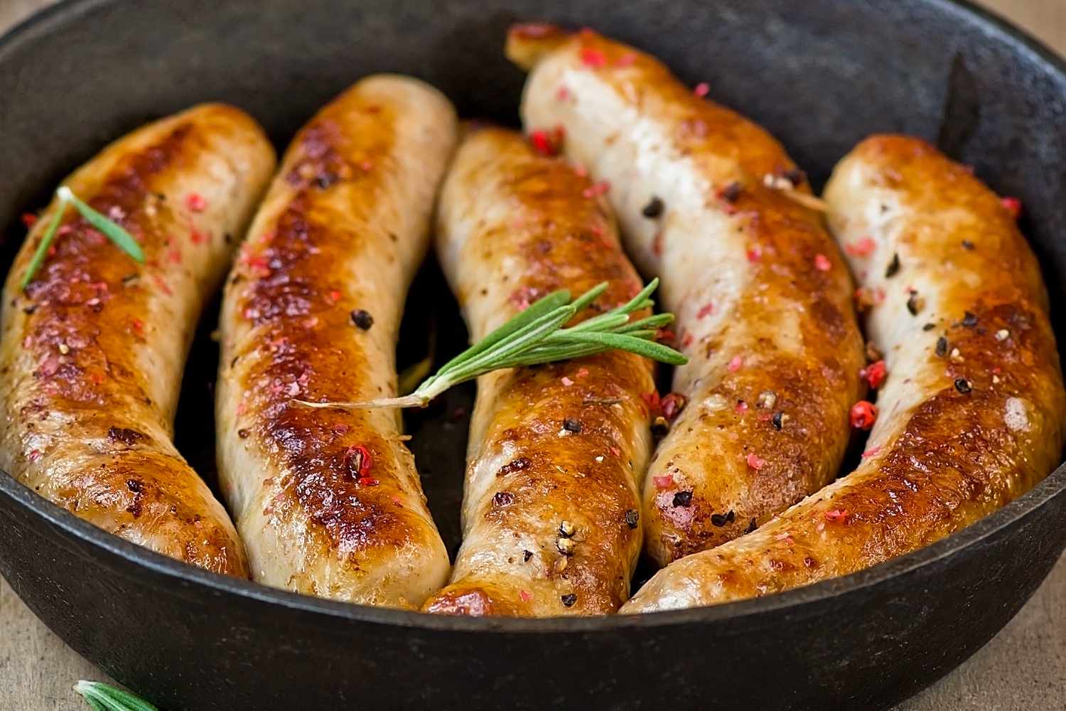 Cooked sausages in a pan garnished with rosemary and peppercorns