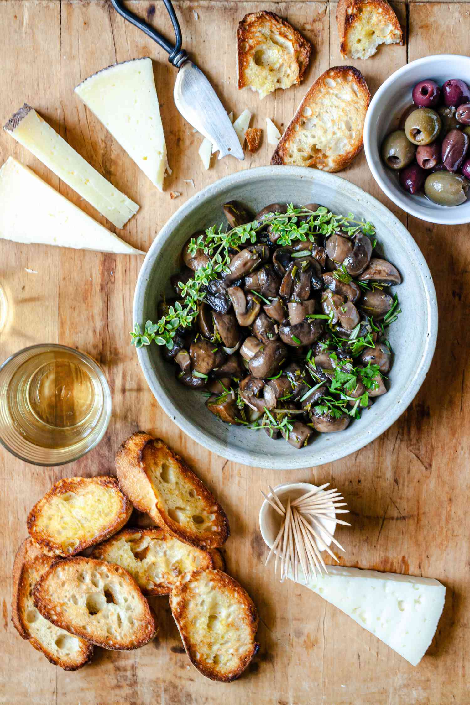 Marinated Mushrooms in a Bowl with Herbs Surrounded by Crostini, Blocks of Cheese, and a Bowl of Olives