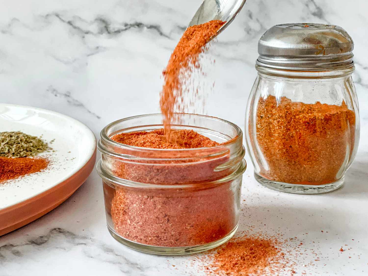 Spoon Pouring Homemade Sazón Mix into a Jar with Shaker in Background