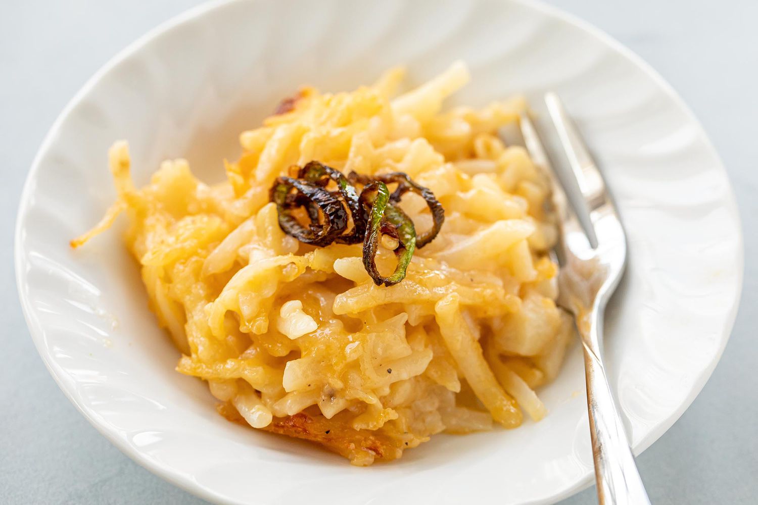 A shallow bowl of potato casserole with crispy serrano chilis on top. A fork is on the right side of the bowl.