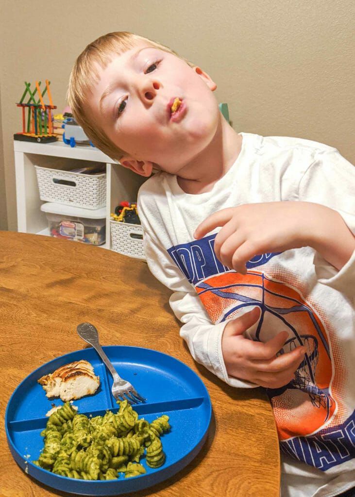 Young boy leaning on table with rotini in his mouth. A blue plate has some more easy kale pesto rotini on one side. A fork rests on another side of the plate next too small pieces of sliced chicken breast.