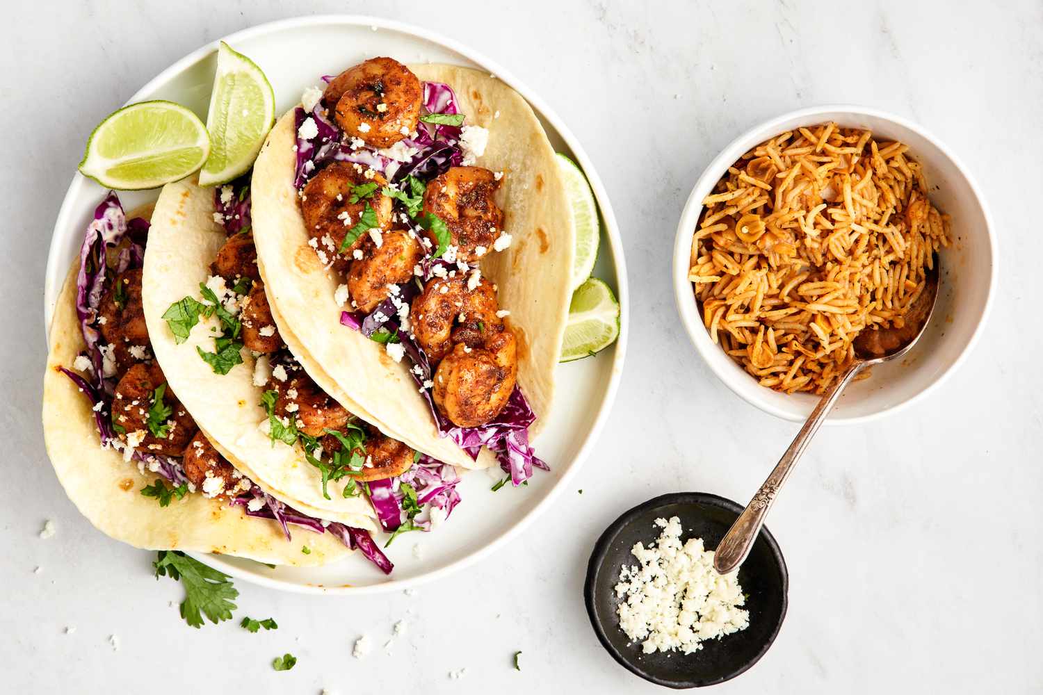 Blackened Shrimp Tacos and Lime Wedges on a Plate Surrounded a Bowl of Rice and a Bowl of Queso Fresco
