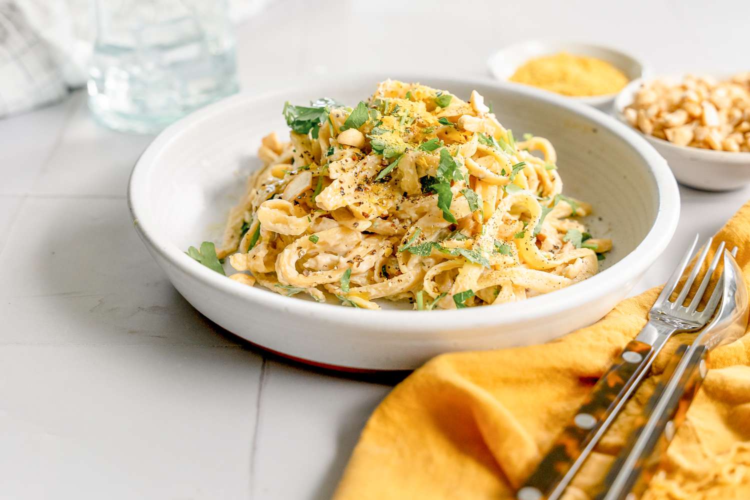 Plate of Super Creamy Vegan Pasta Topped with Parsley, and Next to It, Two Bowls of Ingredients (One: Cashews and Two: Nutritional Yeast), a Glass of Water, and a Utensils on a Table Napkin