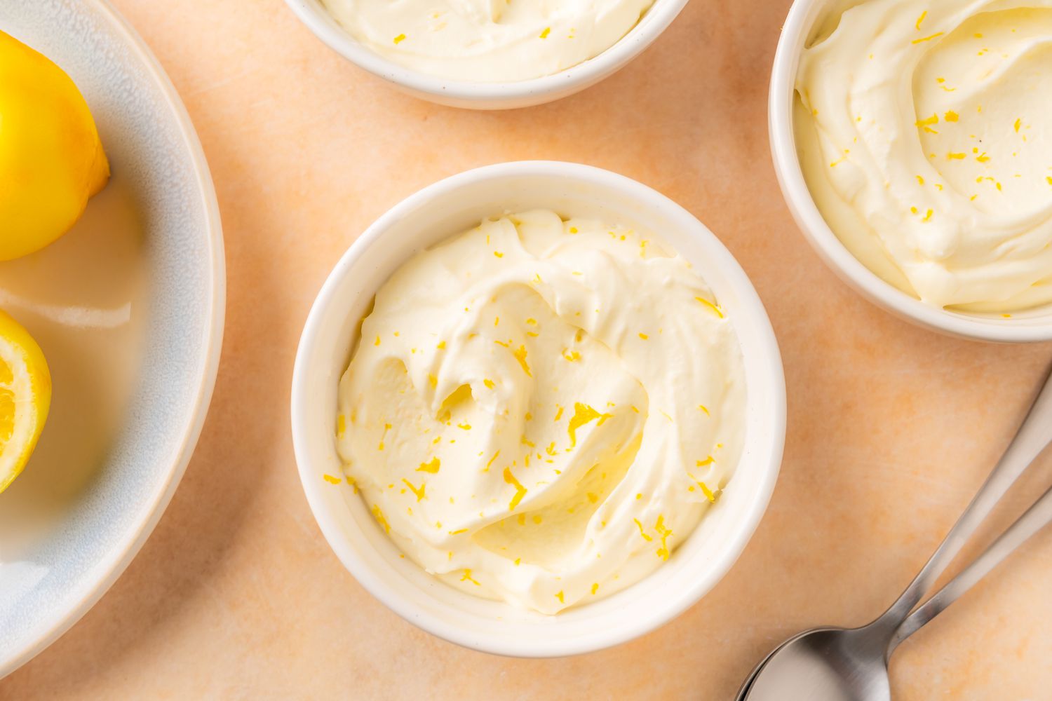 Overhead view of several small white bowls of lemon mousse next to a spoon and plate of lemon slices all on an orange surface