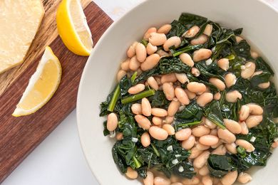Bowl of Italian beans and greens next to a pot with more, a wooden board with lemon wedges, and a block of parmesan