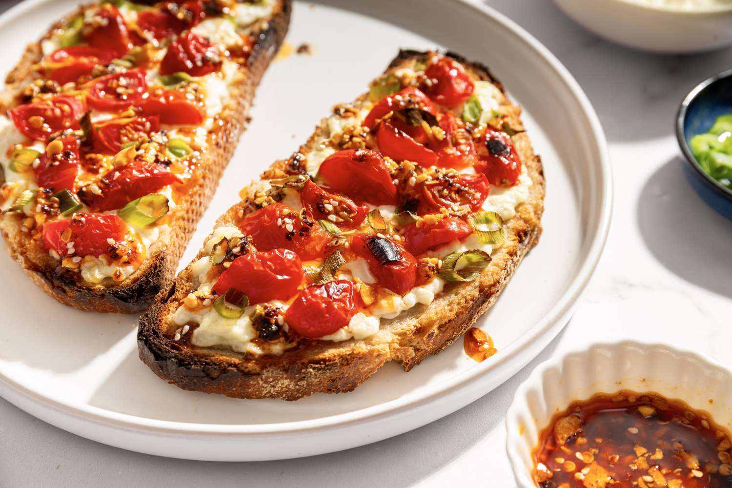 Two slices of toasted bread with cottage cheese roasted tomatoes and seasoning on a white plate served with condiments