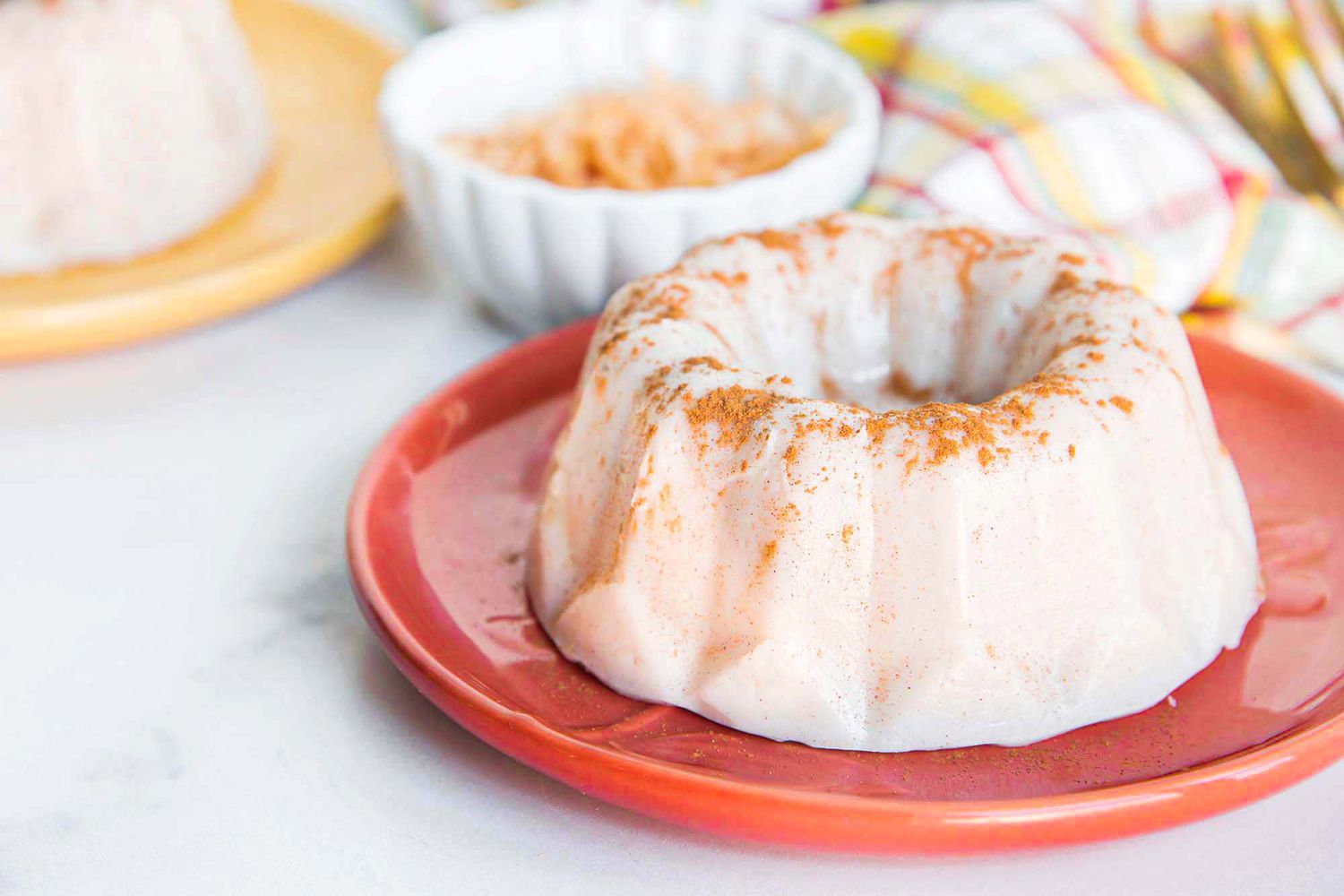 Side view of a coral plate with Puerto Rican panna cotta in the shape of a bundt pan. Ground cinnamon is sprinkled on top. A small white bowl of toasted shredded coconut is behing the plate, as well as a striped dish towel. To the left of the toasted coconut is a light yellow plate with another panna cotta in partial view.