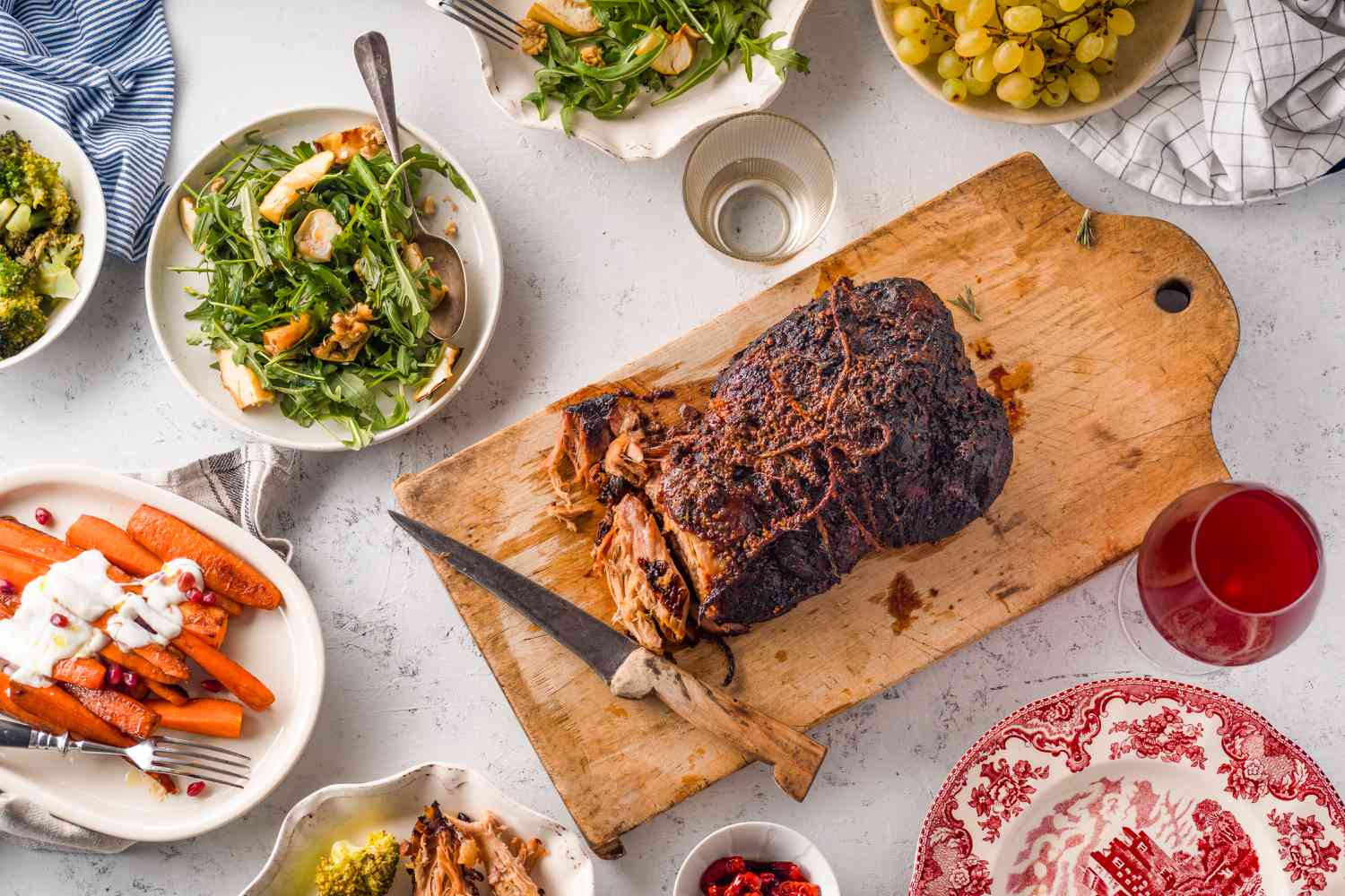 A holiday roast in the center of a festive table surrounded by roasted carrots, an arugula salad, and glasses of wine