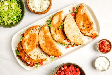 Baked tacos on a platte surrounded by bowls of toppings (bowl of shredded iceberg lettuce, bowl of diced onions, bowl of tomatoes, bowl of salsa, and a bowl of sour cream)