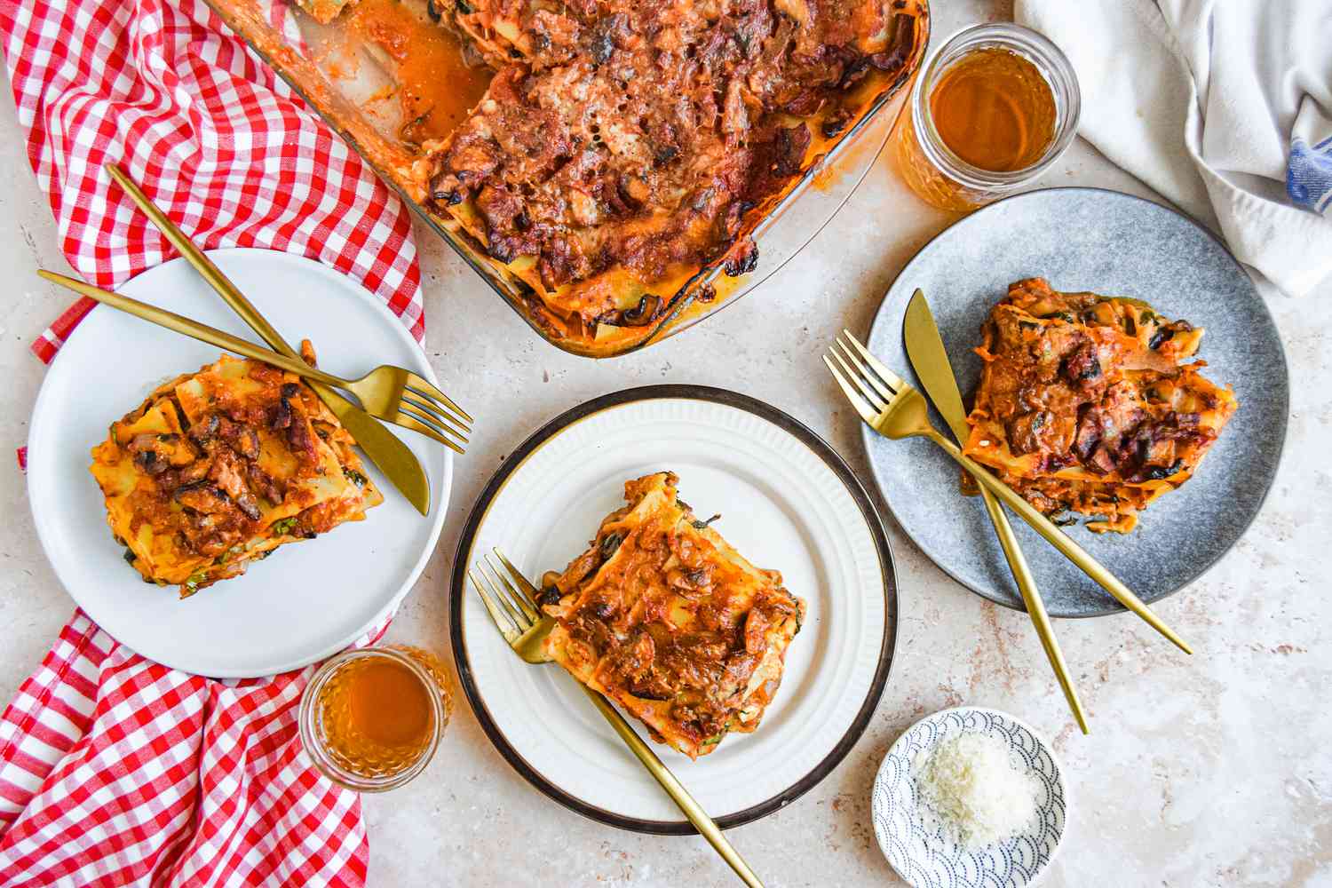 Table Setting: Slices of Vegetarian Spinach and Mushroom Lasagna on Three Plates with Utensils, a Small Saucer of Parmesan, Two Glasses with Drinks, a Checkered Table Cloth, a Table Napkin, and a Casserole Dish with More Lasagna