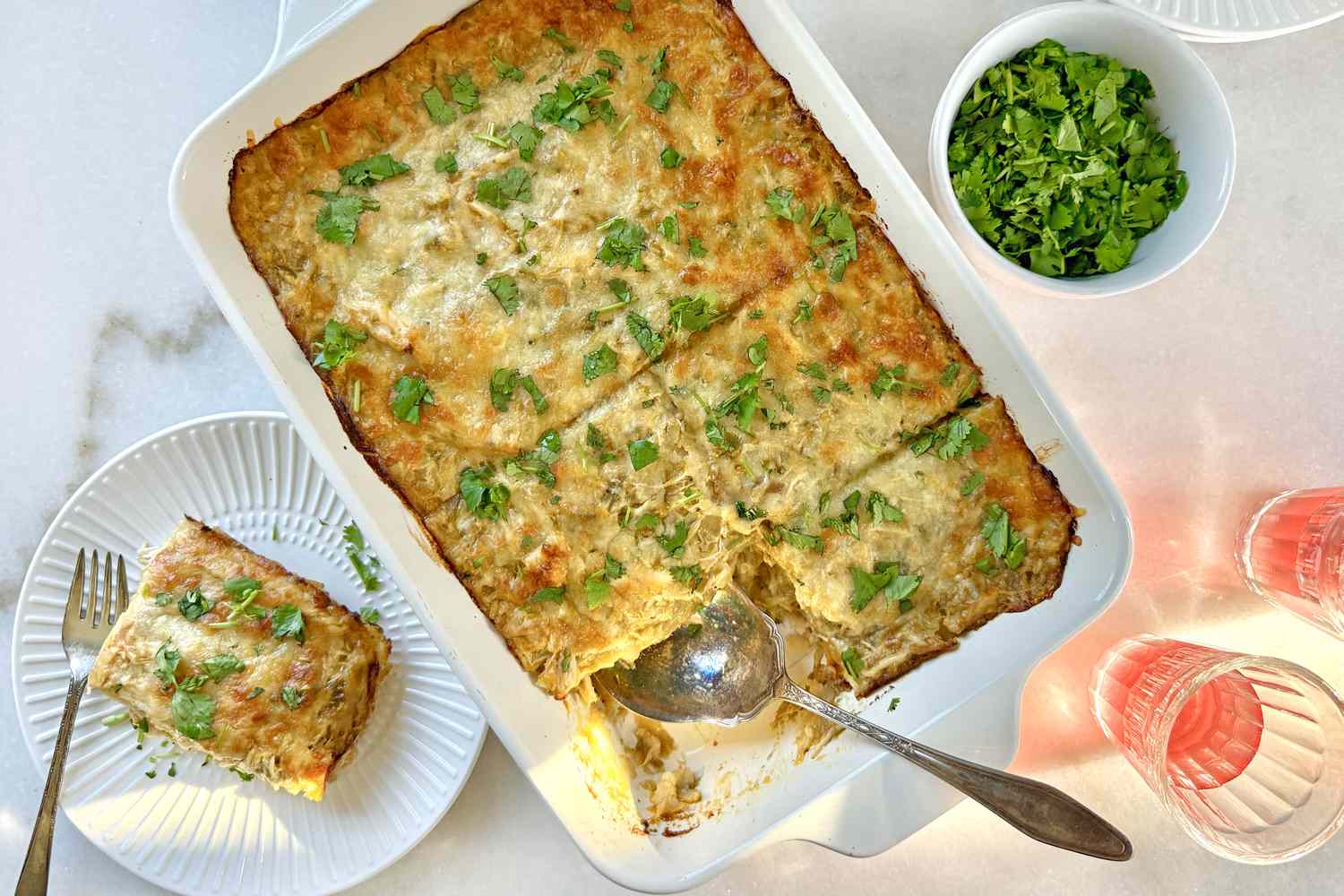 A dish of chicken enchilada casserole with a piece served on a plate garnished with herbs and accompanied by a bowl of fresh garnish