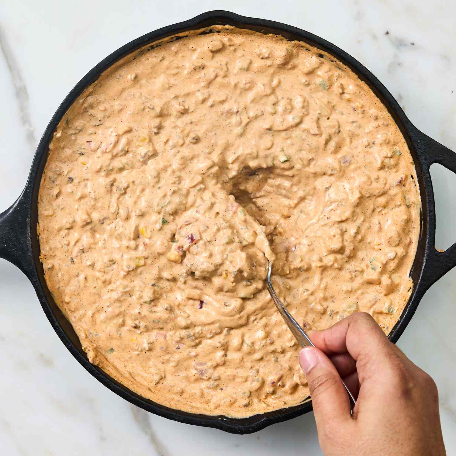 A hand holding a spoon scooping from a skillet containing a creamy dip