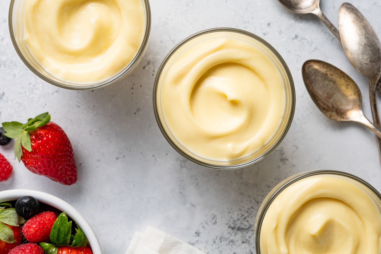 Vanilla Custard in Glass Pudding Jars Surrounded by Berries and Spoons